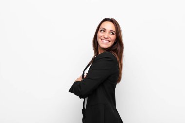 Genç ve güzel iş kadını neşeyle gülümsüyor, mutlu, tatmin olmuş ve rahatlamış hissediyor, kollarını kavuşturup beyaz duvara bakıyor.