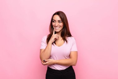Genç ve güzel bir kadın mutlu bir şekilde gülümsüyor ve hayal kuruyor ya da şüphe ediyor, pembe duvara bakıyor.
