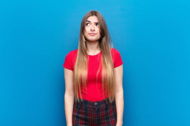 Genç ve güzel bir kadın şaşkın ve şaşkın görünüyor. Bir sorunu çözmeye çalışıyor ya da mavi duvara karşı düşünüyor.