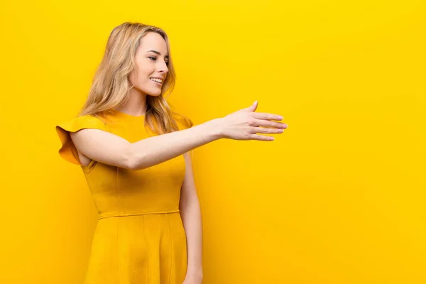 young pretty blonde woman smiling, greeting you and offering a hand shake to close a successful deal, cooperation concept against flat color wall