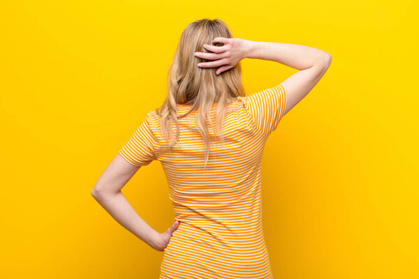 молодая красивая блондинка думает или сомневается, чешет голову, чувствует себя озадаченным и смущенным, спина или задний вид на плоскую стену цвета
