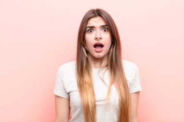 Genç ve güzel bir kadın korkmuş ve şok olmuş hissediyor, ağzı açık pembe duvara karşı şaşkınlık içinde.