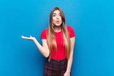 Genç ve güzel bir kadın şaşırmış ve şoka uğramış görünüyor, çenesi düşmüş, mavi duvara karşı eli açık bir obje tutuyor.