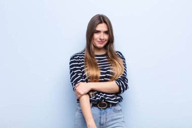 Genç ve güzel bir kadın utangaç ve neşeli bir şekilde gülüyor, dost canlısı ve pozitif ama kendine güveni olmayan bir tavırla yumuşak mavi duvara karşı.