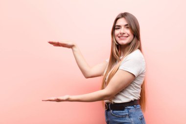 Genç ve güzel bir kadın, iki eli de kenarda olan bir objeyi tutuyor. Fotokopi alanı, pembe duvara karşı bir objeyi gösteriyor, sunuyor ya da reklamını yapıyor.