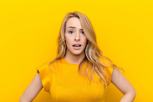 young pretty blonde woman looking very shocked or surprised, staring with open mouth saying wow against flat color wall