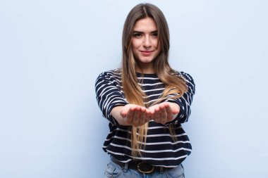 Genç, güzel bir kadın mutlu bir şekilde gülümsüyor. Dost canlısı, kendine güvenen, pozitif bakışlı, yumuşak mavi duvara karşı bir nesne ya da konsept sunuyor ve gösteriyor.