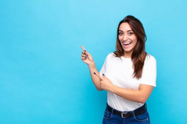 Genç, güzel bir kadın sevinçli ve şaşırmış hissediyor. Şok olmuş bir ifadeyle gülümsüyor ve mavi duvara doğru işaret ediyor.
