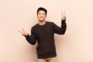 Genç Çinli adam gülümsüyor ve mutlu görünüyor, arkadaş canlısı ve memnun, iki eliyle düz duvara karşı zafer ya da barış işareti yapıyor