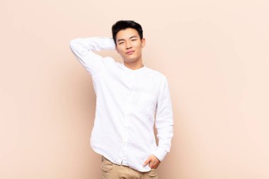 Genç Çinli adam neşeli ve tesadüfen gülümsüyor, pozitif, mutlu ve kendine güvenen bir bakışla düz renkli duvara bakıyor.