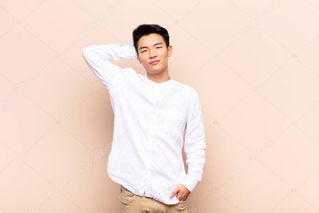 young chinese man smiling cheerfully and casually, taking hand to head with a positive, happy and confident look against flat color wall