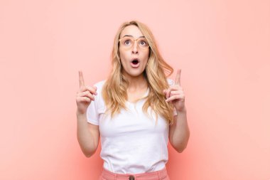 Genç, güzel, sarışın bir kadın huşu içinde ve ağzı açık bir şekilde yukarı bakıyor. Düz renkli duvara doğru şaşkın ve şaşkın bir şekilde bakıyor.