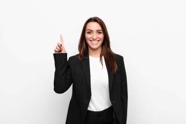 Genç ve güzel iş kadını mutlu bir şekilde gülümsüyor, tek eliyle beyaz duvara uzayı gösteriyor.