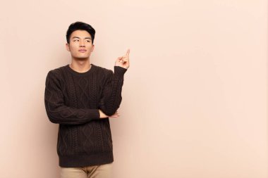 Genç Çinli adam mutlu bir şekilde gülümsüyor ve yan bakıyor, merak ediyor, düşünüyor ya da düz renkli duvara karşı bir fikri var.