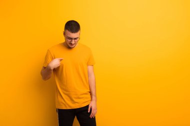 Genç yakışıklı adam neşeyle ve gelişigüzel gülümsüyor, aşağıya bakıyor ve düz arkaplana karşı göğsüne işaret ediyor.