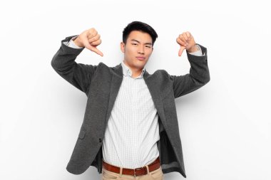 Genç Çinli adam üzgün, hayal kırıklığına uğramış ya da kızgın görünüyor. Anlaşmazlıkta baş parmağını gösteriyor. Düz renkli duvara karşı hayal kırıklığına uğramış hissediyor.