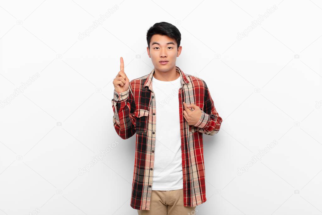 young chinese man feeling proud and surprised, pointing to self confidently, feeling like successful number one against flat color wall