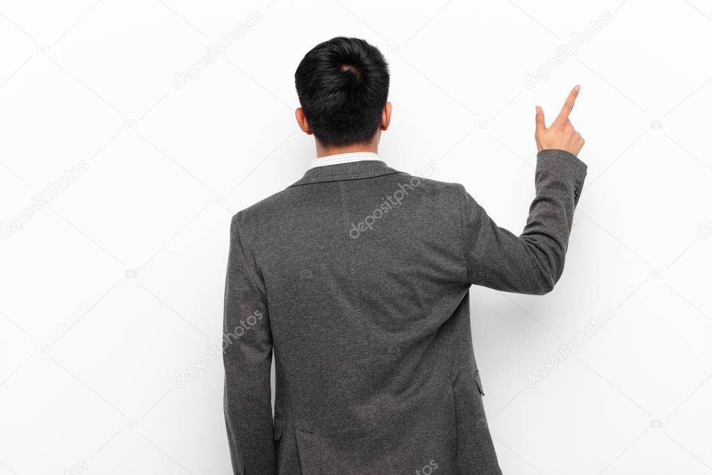 young chinese man standing and pointing to object on copy space, rear view against flat color wall