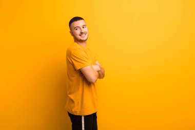 Genç yakışıklı adam neşeyle gülümsüyor, mutlu, tatmin olmuş ve rahatlamış, kollarını kavuşturup düz arkaplana bakıyor.