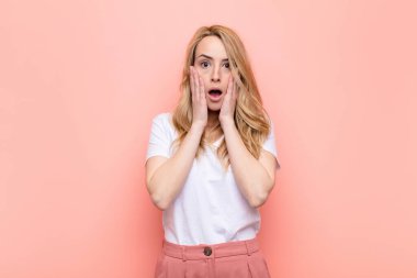 Genç ve güzel sarışın kadın şok olmuş ve korkmuş hissediyor, ağzı açık ve elleri yanaklarında düz renkli duvara bakıyor.