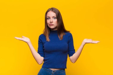 Genç kızıl saçlı kadın şaşkın ve şaşkın hissediyor, şüpheci, farklı seçenekleri tartıyor ya da seçiyor düz duvara karşı komik bir ifadeyle