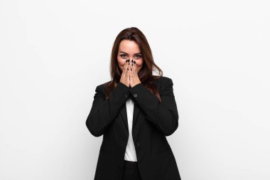 Genç ve güzel iş kadını mutlu, neşeli, şanslı ve şaşkın görünüyor. İki eli de beyaz duvara dayalı ağzını kapatıyor.