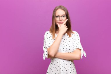 Genç kızıl saçlı kadın ciddi, düşünceli ve güvensiz görünüyor, bir kolu çapraz ve çene üzerinde, düz duvara karşı seçenekleri tartıyor