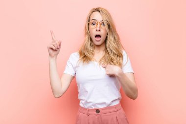 Genç ve güzel sarışın kadın gururlu ve şaşırmış hissediyor, kendine güvenen, düz renkli duvara karşı bir numara gibi hisseden.