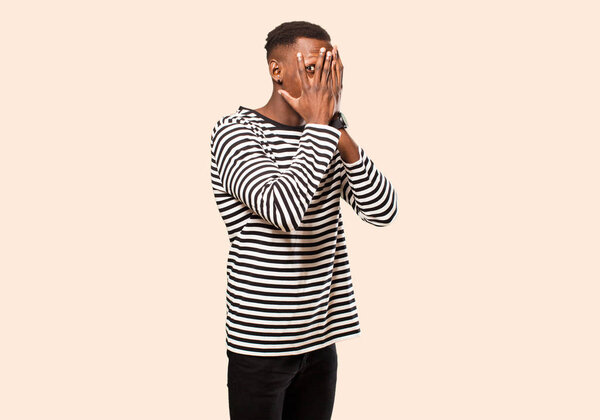 Young african american black man covering face with hands, peeking between fingers with surprised expression and looking to the side against beige wall
