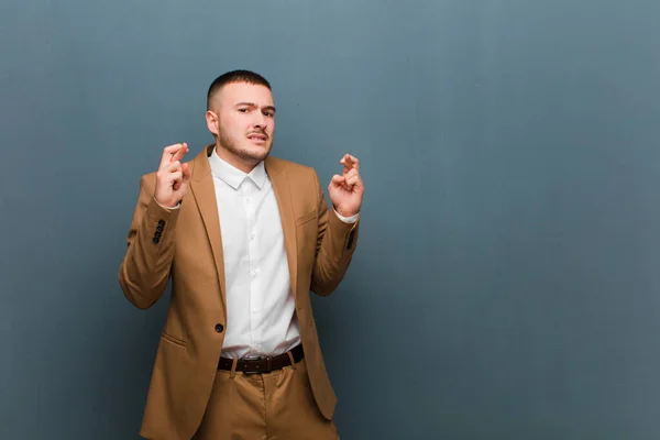 young handsome businessman crossing fingers anxiously and hoping for good luck with a worried look against flat background