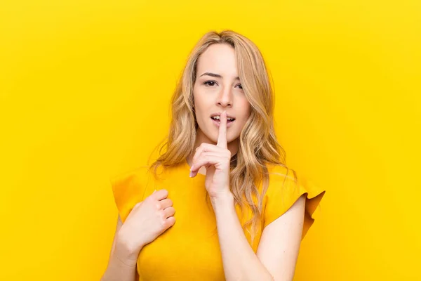 young pretty blonde woman looking serious and cross with finger pressed to lips demanding silence or quiet, keeping a secret against flat color wall