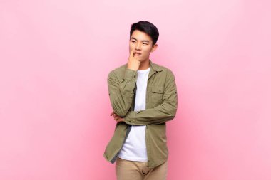 Şaşırmış, gergin, endişeli ya da korkmuş bakışları olan genç Çinli bir adam düz renkli duvara karşı fotokopi odasına doğru bakıyor.