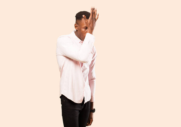 Young african american black man raising palm to forehead thinking oops, after making a stupid mistake or remembering, feeling dumb against beige wall
