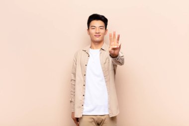 Genç Çinli adam gülümsüyor ve arkadaş canlısı görünüyor. Üç ya da üçüncüyü gösteriyor. Elleri önde, düz renkli duvara doğru sayıyor.