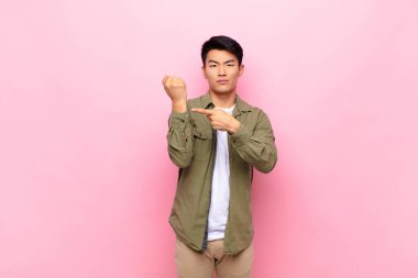 Genç Çinli adam sabırsız ve kızgın görünüyor, saati gösteriyor, dakiklik istiyor, düz renkli duvara karşı zamanında olmak istiyor.