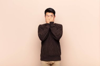 Genç Çinli adam elleriyle yüzünü kapatıyor, parmaklarının arasından şaşkın bir ifadeyle bakıyor ve düz renkli duvara bakıyor.
