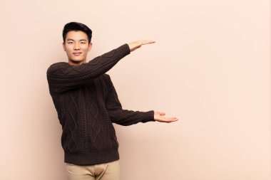 Genç Çinli adam gülümsüyor, mutlu, pozitif ve tatmin olmuş hissediyor, düz renkli duvara karşı fotokopi alanında nesne ya da konsept tutuyor ya da gösteriyor