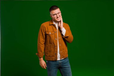 Genç yakışıklı adam yanağını tutuyor ve acı çekiyor, hasta, sefil ve mutsuz hissediyor, düz arkaplanda bir dişçi arıyor.