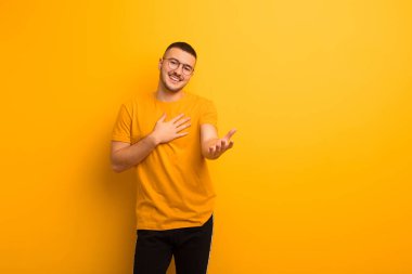 Genç yakışıklı adam mutlu ve aşık, bir eli kalbinin yanında gülümsüyor ve diğer eli düz arkaplana doğru uzanıyor.