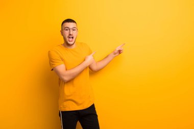 Genç yakışıklı adam mutlu ve şaşırmış hissediyor, şok olmuş bir ifadeyle gülümsüyor ve düz arkaplanı işaret ediyor.
