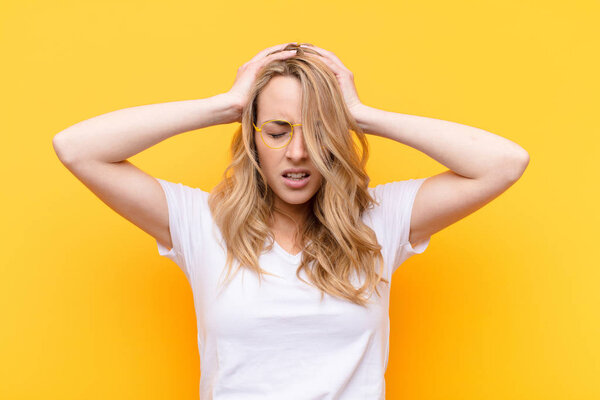 young pretty blonde woman feeling stressed and frustrated, raising hands to head, feeling tired, unhappy and with migraine against flat color wall