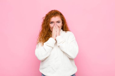 Genç kızıl saçlı kadın mutlu, neşeli, şanslı ve şaşkın görünüyor. İki eli de pembe duvarda.