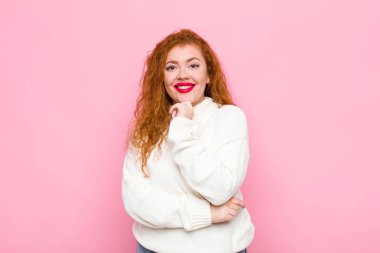 Genç kızıl saçlı kadın mutlu görünüyor ve gülümsüyor. Elleri çenesinde, merak ediyor ya da soru soruyor. Pembe duvarla seçenekleri karşılaştırıyor.