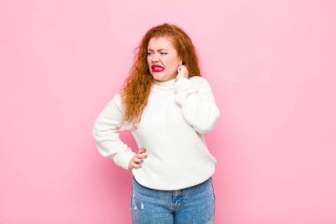 Genç kızıl saçlı kadın stresli, hayal kırıklığına uğramış ve yorgun hissediyor. Pembe duvara karşı endişeli ve sorunlu bir bakışla boynunu ovalıyor.