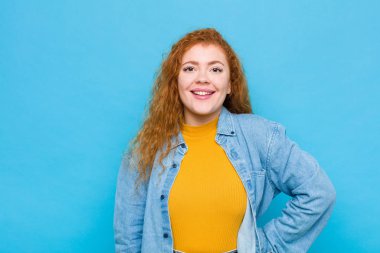 Genç kızıl saçlı kadın mutlu bir şekilde gülümsüyor, eli kalçasında ve kendinden emin, pozitif, gururlu ve dost canlısı bir tavırla mavi duvara karşı.