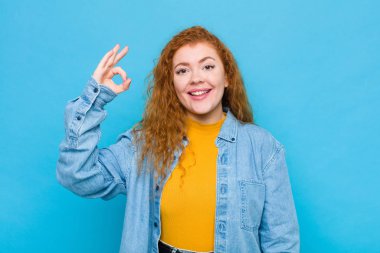 Genç kızıl saçlı kadın kendini mutlu, rahat ve tatmin olmuş hissediyor. Onayını iyi bir hareketle gösteriyor. Mavi duvara gülümsüyor.