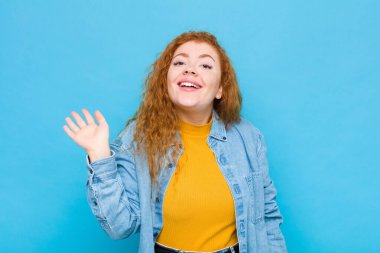 Genç kızıl saçlı kadın mutlu ve neşeyle gülümsüyor, el sallıyor, seni selamlıyor, ya da mavi duvara veda ediyor.