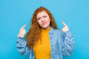 Kötü tavırlı genç kızıl saçlı kadın gururlu ve agresif görünüyor, yukarı bakıyor ya da mavi duvara yaslanmış ellerle dalga geçiyor.