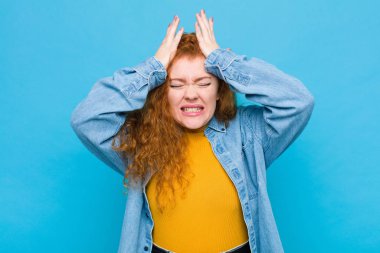 Genç kızıl saçlı kadın stresli ve endişeli, depresif ve hayal kırıklığına uğramış hissediyor, iki elini de mavi duvara doğru kaldırıyor.