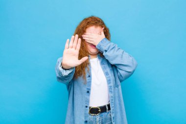 Genç kızıl saçlı kadın yüzünü eliyle kapatıyor ve kamerayı durdurmak için diğer elini öne koyuyor. Mavi duvara karşı fotoğraf ya da resim çekmeyi reddediyor.